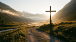A large wooden cross overlooks a misty mountain valley with a winding dirt road at sunrise.