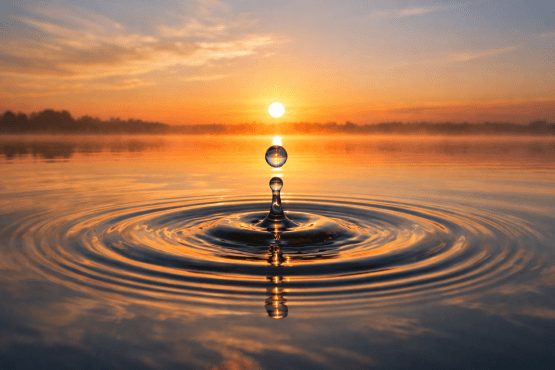 Water drop suspended above lake surface reflecting sunrise with ripples around it