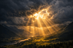 Sunlight rays shining through dark clouds onto a valley with a river and mountains