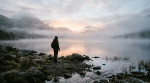 Person with backpack standing on rocky shore looking at misty lake and mountains at sunrise